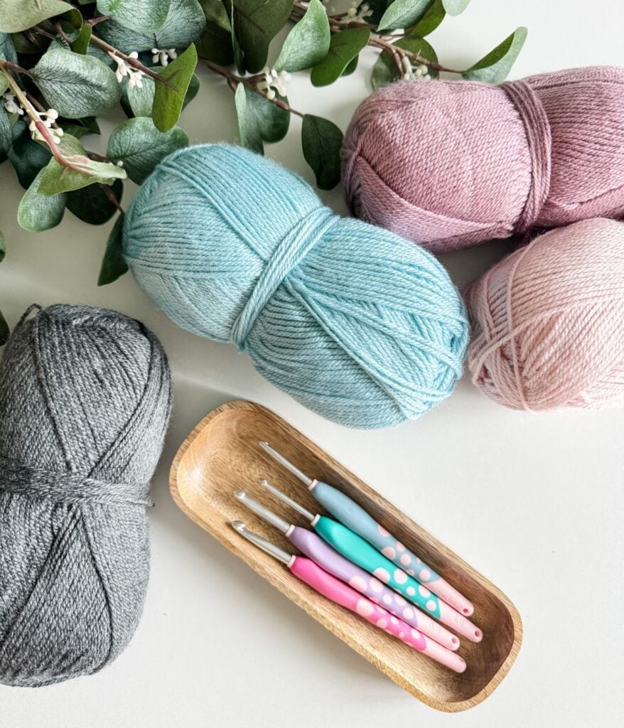 A flat lay of knitting supplies including pastel-colored yarn balls, granny square crochet hooks in a wooden bowl, and green eucalyptus branches on a white surface.