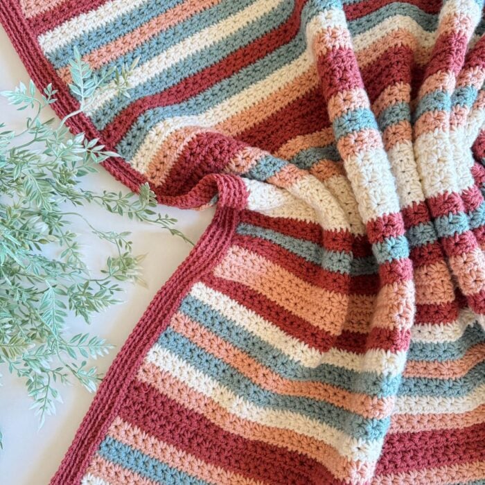Handcrafted striped crochet pullover with pastel colors alongside a sprig of greenery on a white surface.