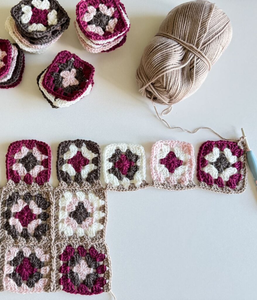 Crocheted granny squares in beige, white, and pink arranged next to yarn and a crochet hook on a light surface.