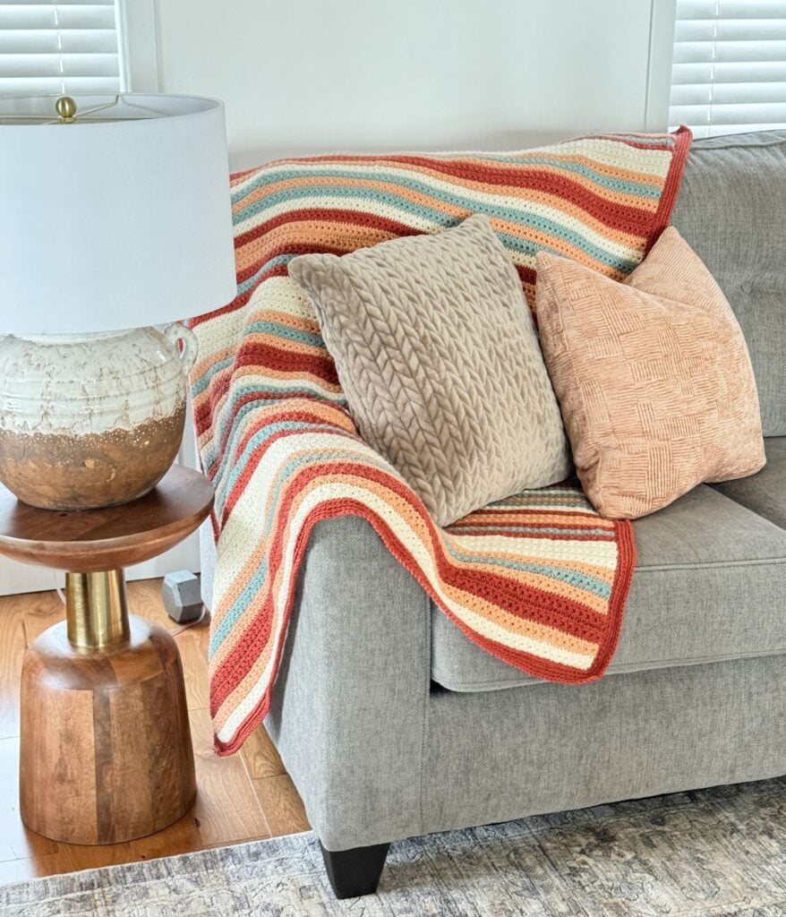 A cozy living room corner with a grey sofa, decorative cushions, an easiest children's crochet pullover as a colorful knitted throw, and a wooden side table with a lamp.
