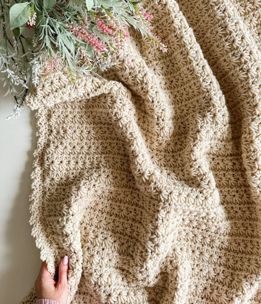 A hand holding a corner of a textured beige children's crochet pullover next to a bouquet of flowers.