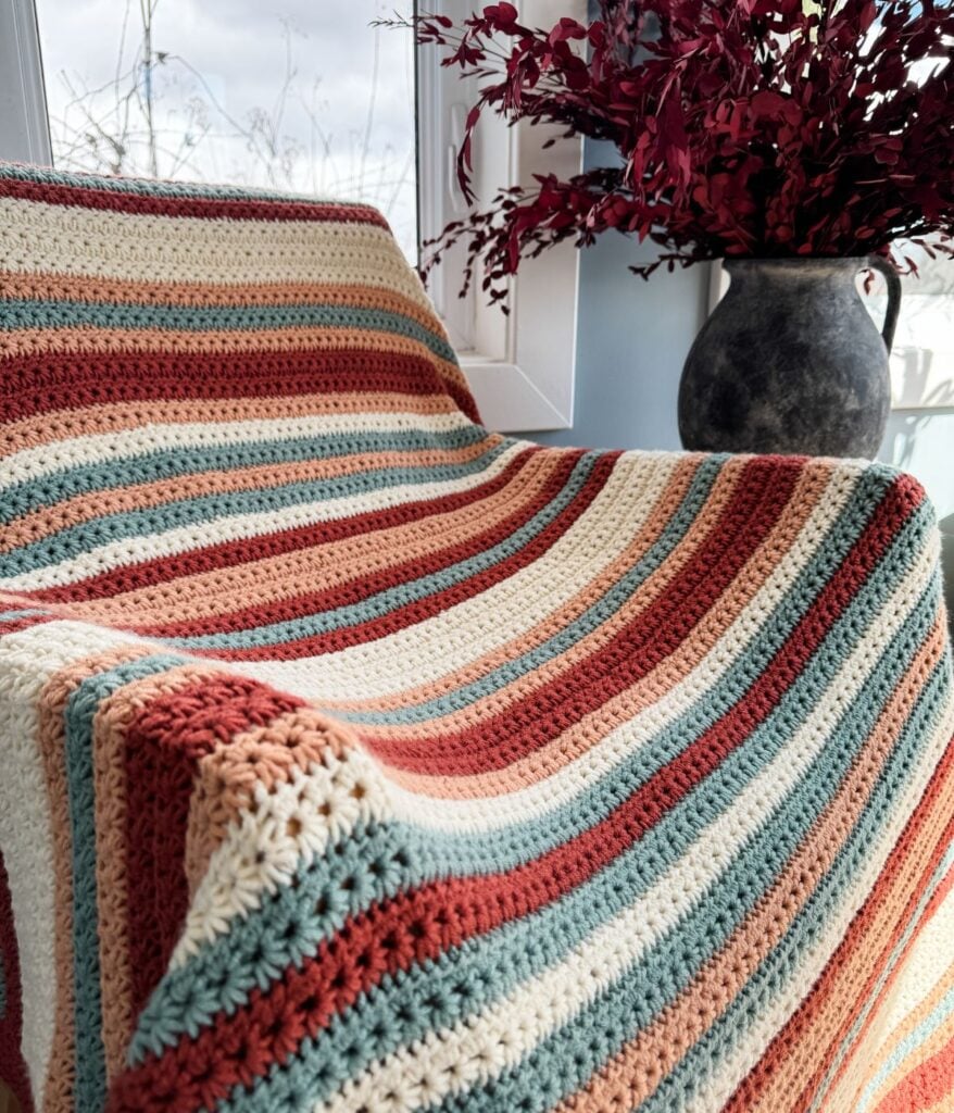 Crochet striped blanket draped over a chair near a window with a vase of red leaves.