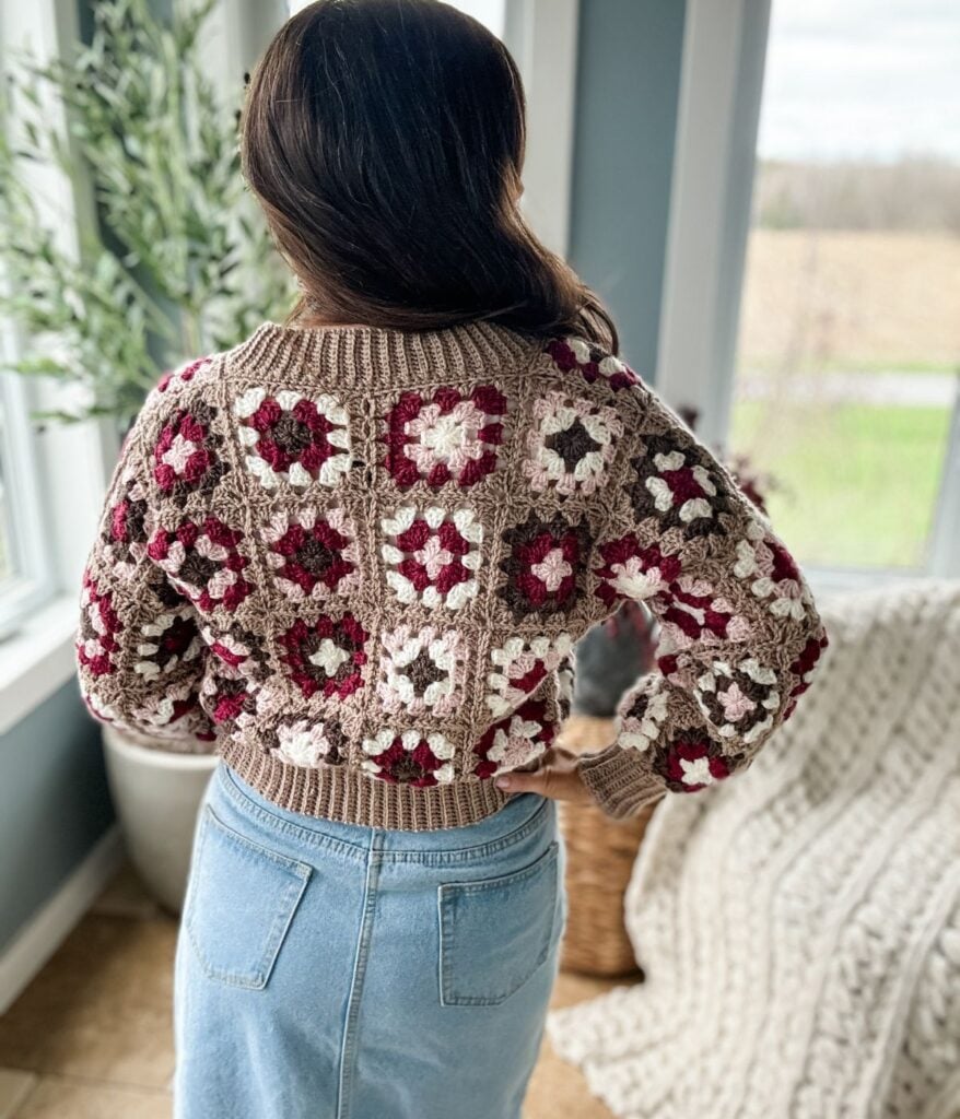 Woman standing by a window, viewed from behind, wearing a granny square patterned sweater and blue jeans, looking out at a field.