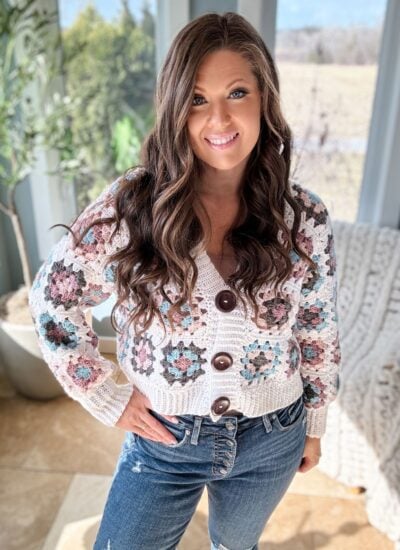 A woman with long, wavy hair smiling indoors, wearing a granny square crochet sweater and jeans, with a bright window and greenery behind her.