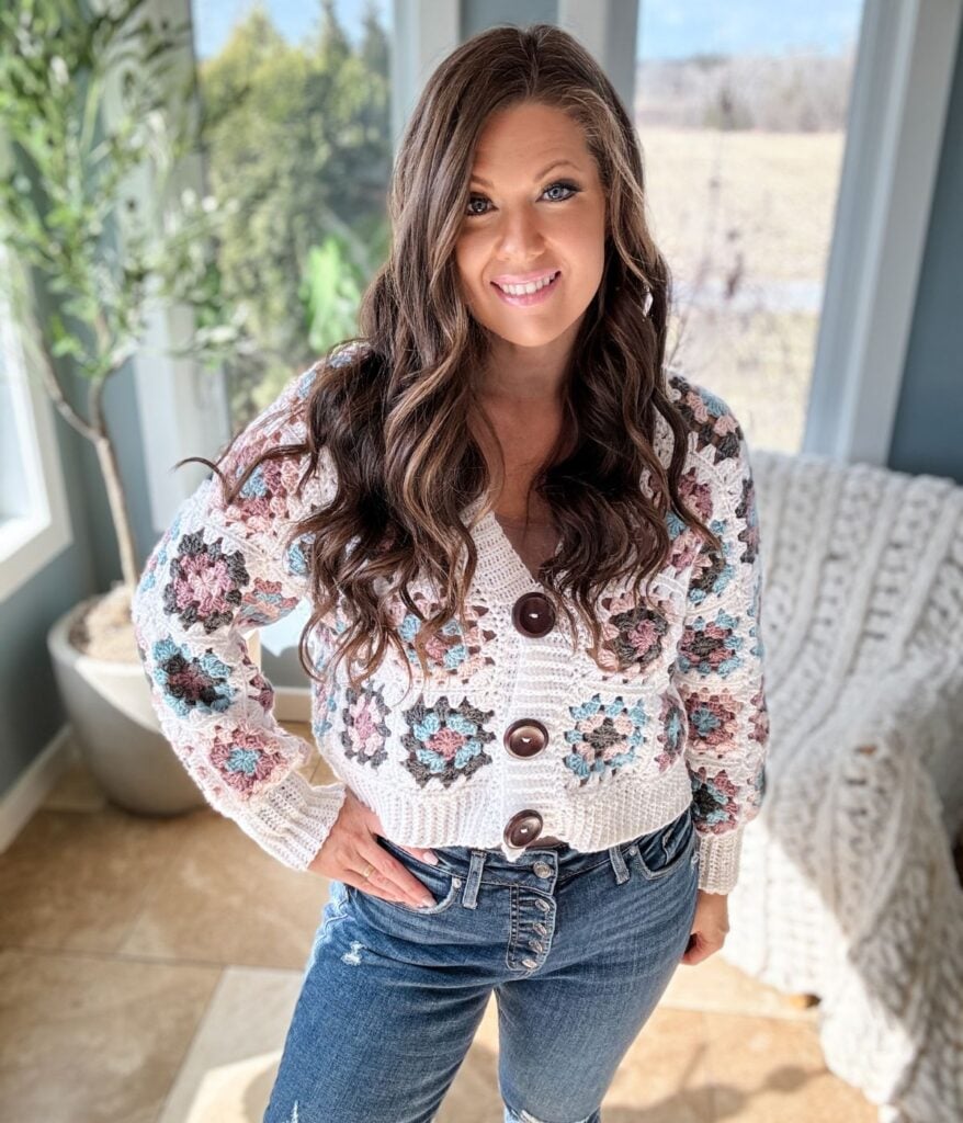 A woman with long, wavy hair smiling indoors, wearing a granny square crochet sweater and jeans, with a bright window and greenery behind her.