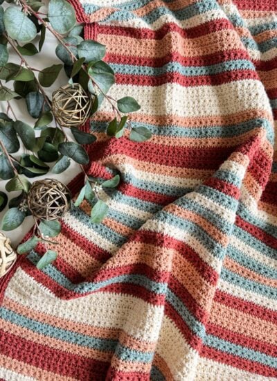 A handmade striped crochet blanket in warm tones, designed as the easiest children's pullover, alongside decorative green eucalyptus branches and wooden spheres on a white surface.