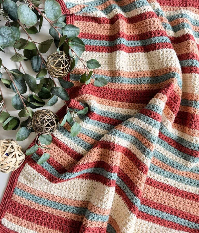 A handmade striped crochet blanket in warm tones, designed as the easiest children's pullover, alongside decorative green eucalyptus branches and wooden spheres on a white surface.