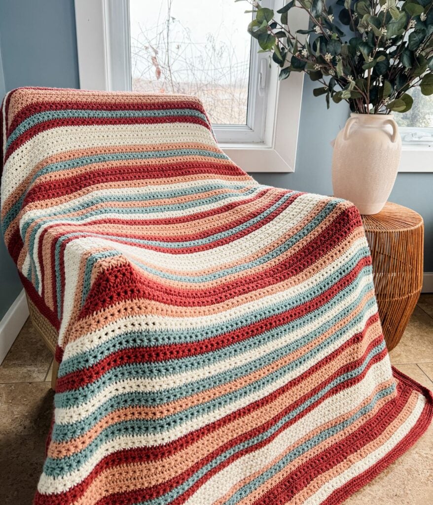 A handcrafted striped crochet blanket is draped over a chair by a window.