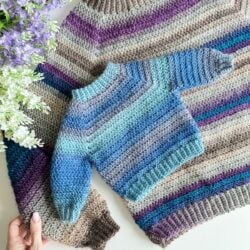 A handcrafted crochet cardigan laid out flat next to purple flowers.