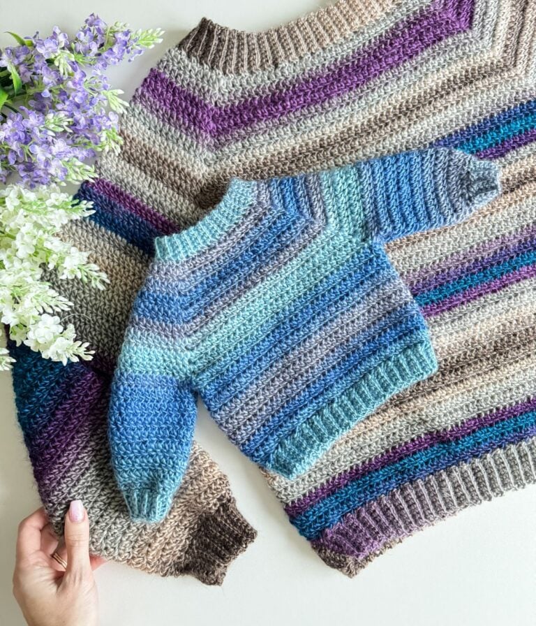 A handcrafted crochet cardigan laid out flat next to purple flowers.