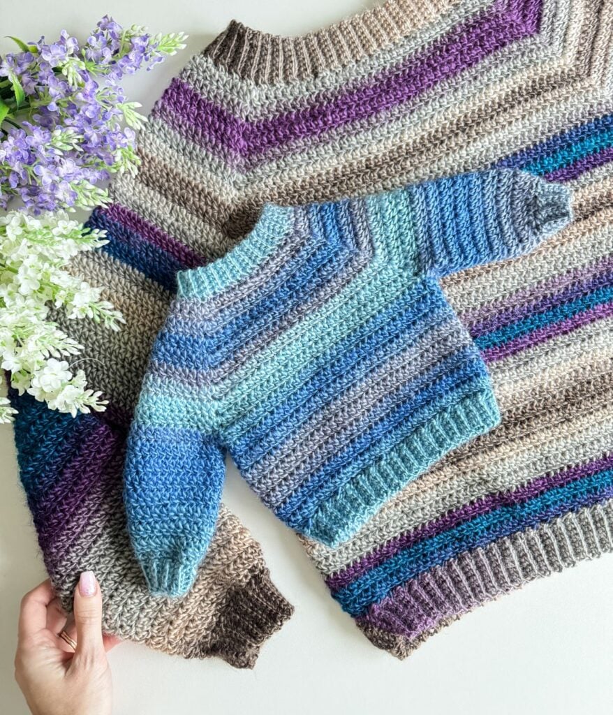 A handcrafted crochet cardigan laid out flat next to purple flowers.