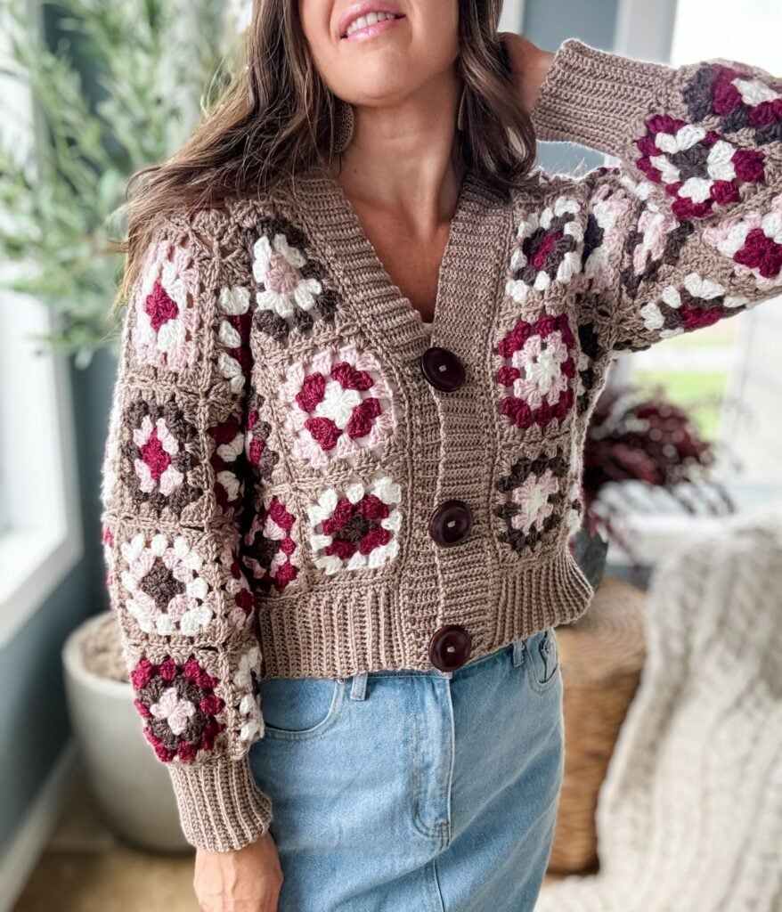 Woman wearing a granny square crochet cardigan in beige, pink, and white, paired with blue jeans, standing indoors.