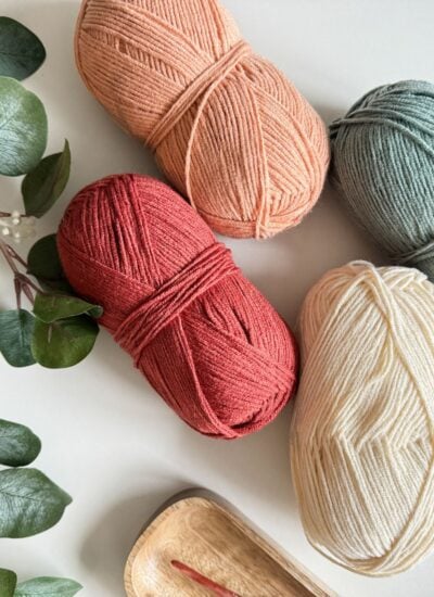 Four skeins of yarn in various colors for the easiest children's crochet pullover, with knitting needles and green leaves on a light background.