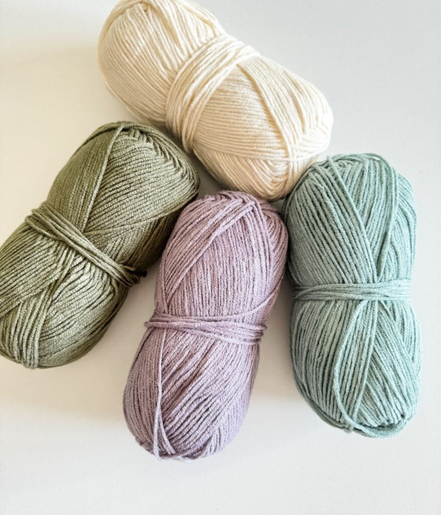 Four skeins of yarn in pastel green, lavender, cream, and sage colors arranged on a white surface.