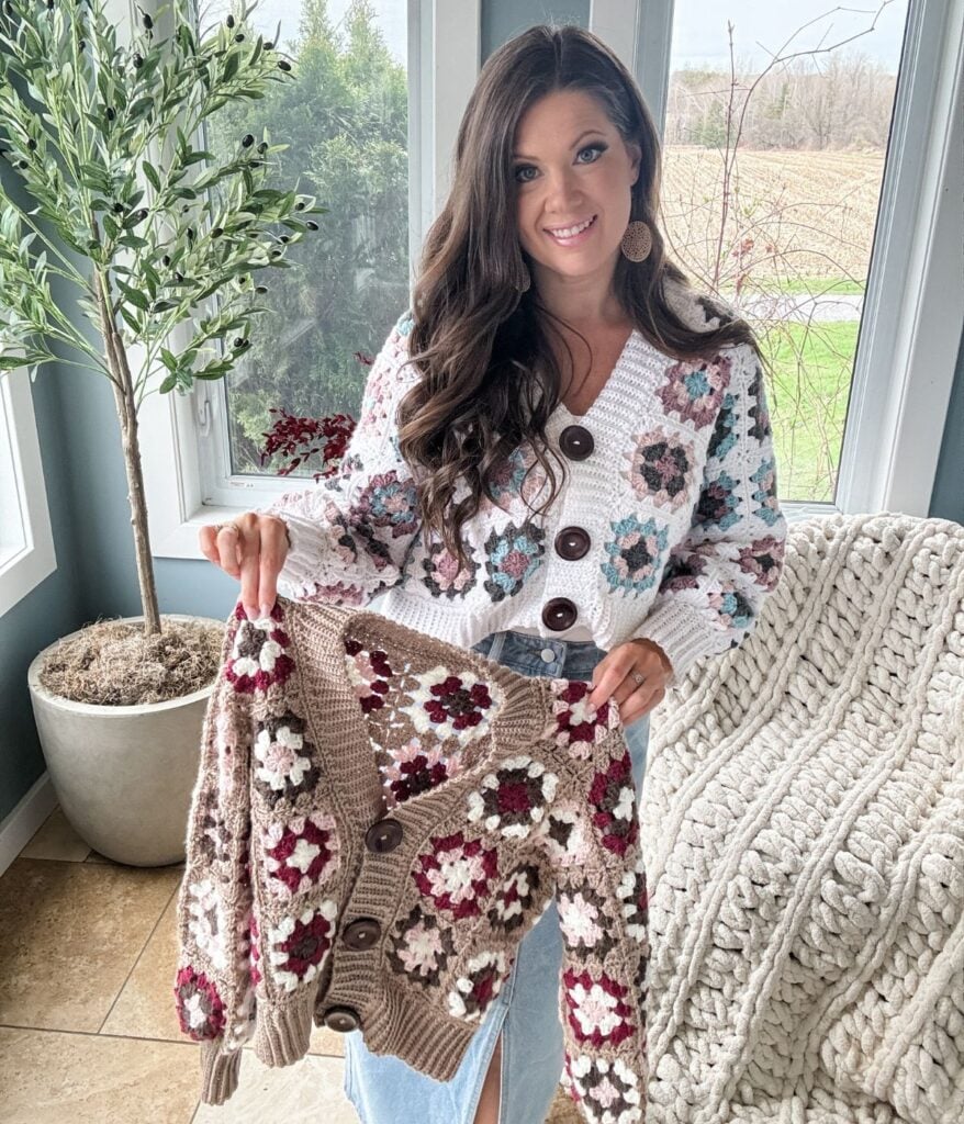 A woman smiling, holding a knitted sweater with granny square patterns, standing indoors beside a window and a potted plant.