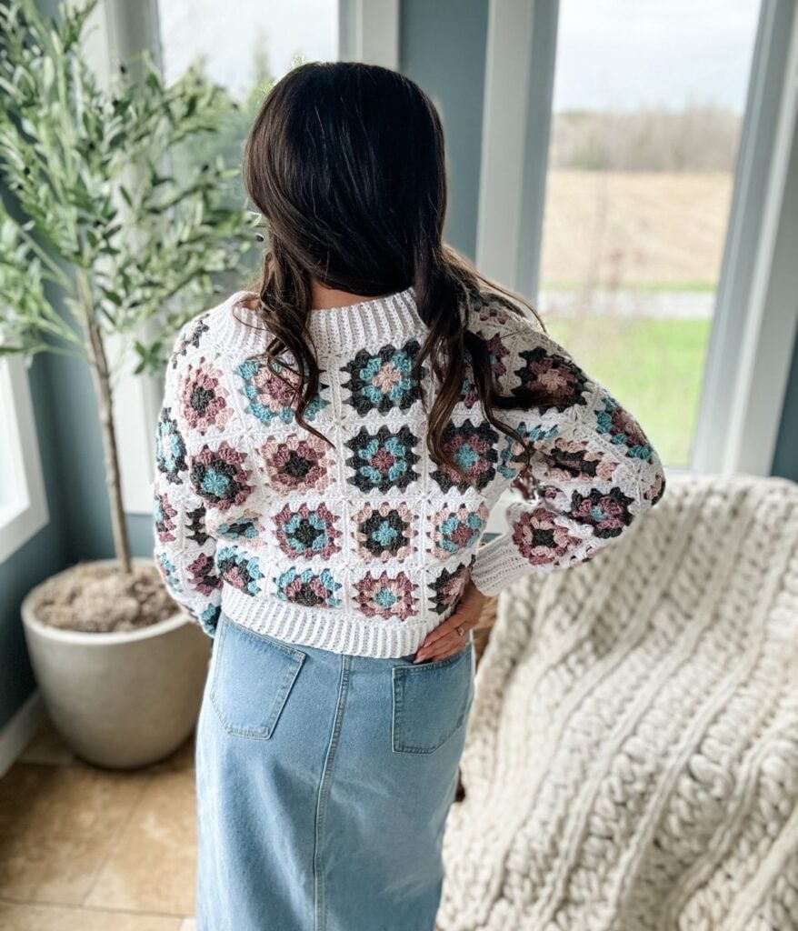 Woman standing by a window, wearing a granny square crochet sweater and denim skirt, looking out onto a field.