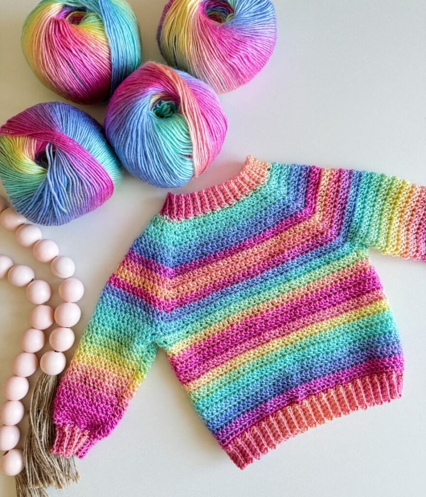 A colorful crochet rainbow cardigan alongside vibrant yarn skeins and pink beads.
