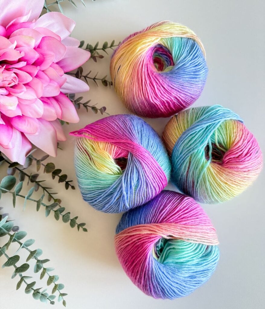 Multicolored yarn balls for a crochet cardigan alongside a pink artificial flower and greenery on a white surface.