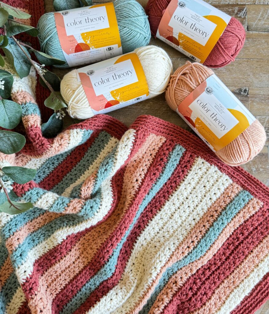A Crochet Striped Blanket in red, cream, pink, and green is displayed next to four skeins of coordinating Color Theory yarn on a wooden surface with some greenery.