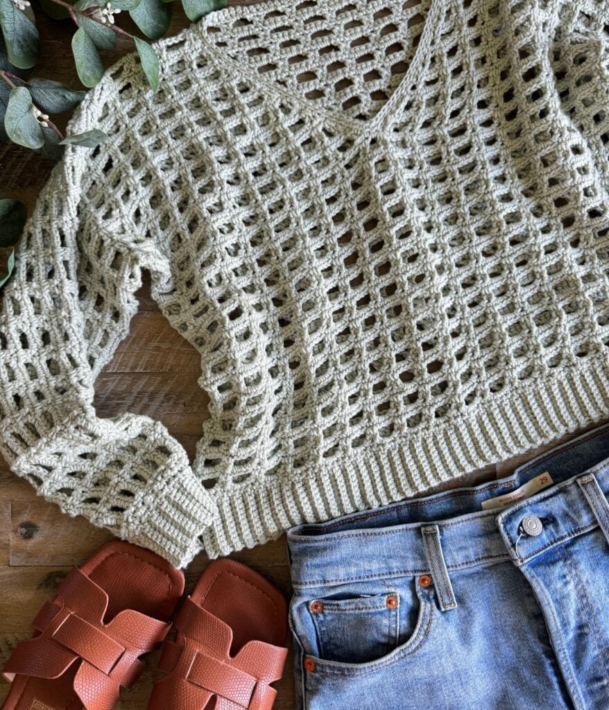 A beige crochet mesh top is laid out next to blue jeans and a pair of red sandals on a wooden surface with green leaves nearby.