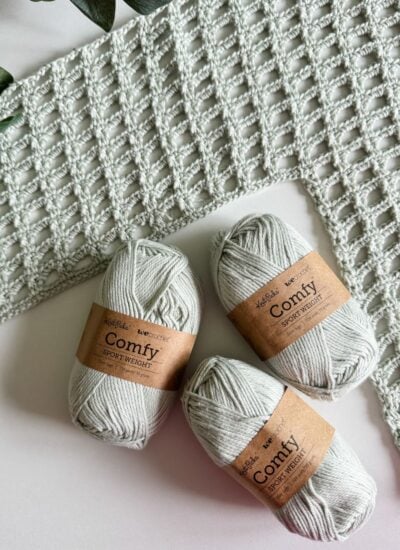 Three skeins of beige yarn labeled "Comfy Sport Weight" are placed next to a partially crocheted mesh top, with green foliage in the background.