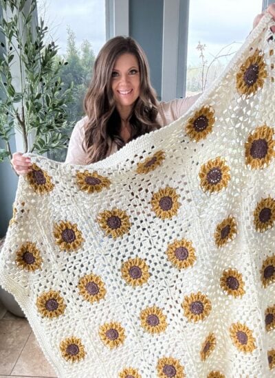 A woman smiling, holding up a crocheted beige blanket with yellow and brown flower patterns, standing indoors by windows and greenery.