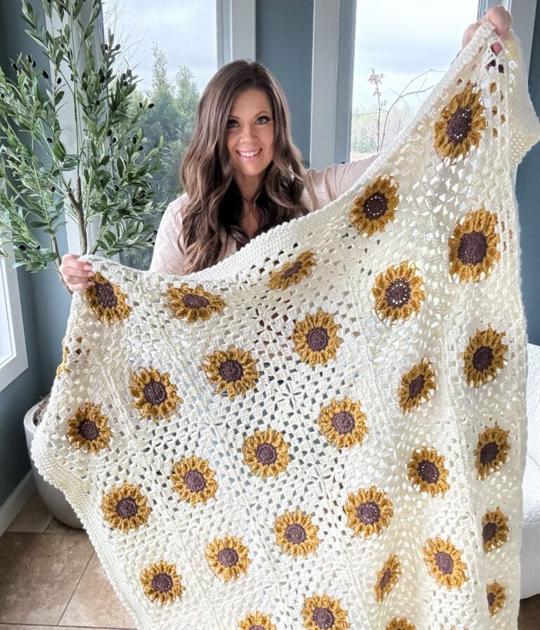 A woman smiling, holding up a crocheted beige blanket with yellow and brown flower patterns, standing indoors by windows and greenery.