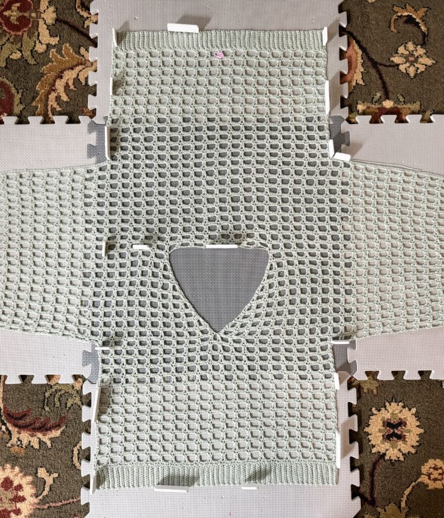 A crocheted mesh top in a light green color is laid flat on a patterned mat, showcasing the front, back, and sleeves. The top has a hole at the center for the neck and is partially completed.