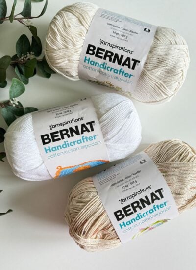 Three skeins of bernat handicrafter cotton yarn, two in white and one in variegated beige and white, arranged on a light surface with green foliage in the background, ideal for a summer crochet purse pattern.