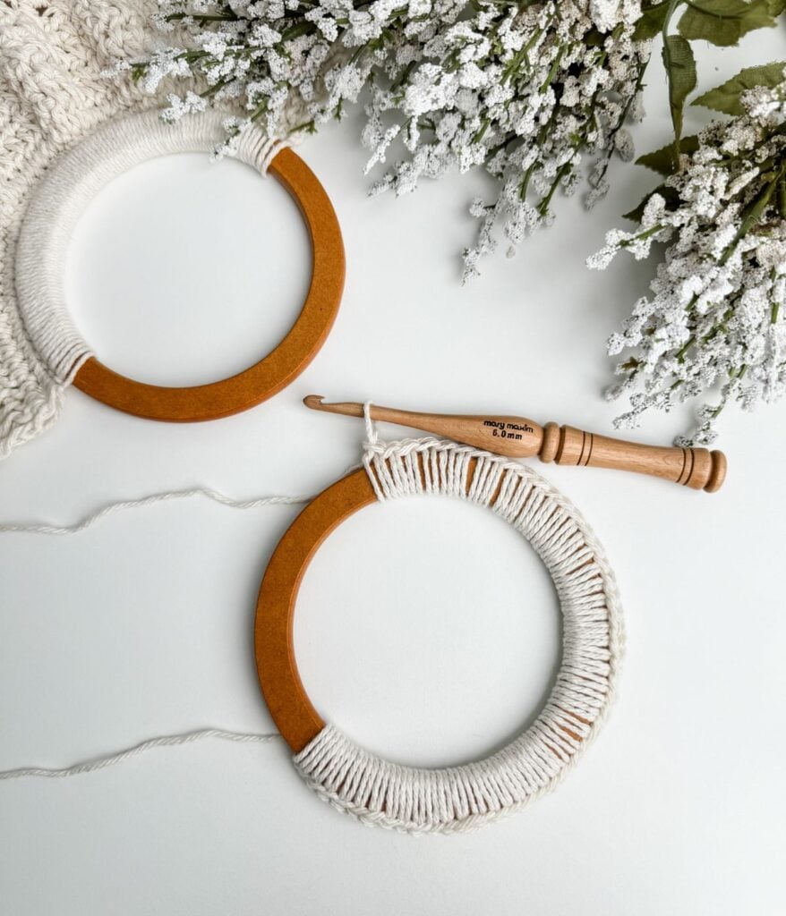 Two wooden macrame rings with white thread and a crochet hook, next to white flowers on a summer crochet pattern background.