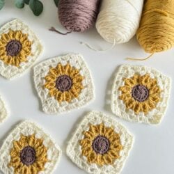 Crocheted sunflower squares in white, gold, and brown, alongside yarn skeins in pink, white, and mustard colors, arranged on a white surface.