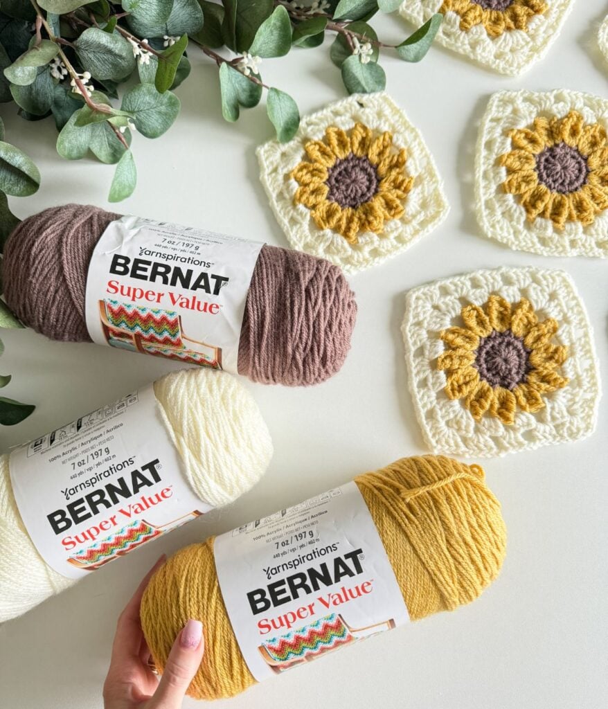 A person's hand arranging three skeins of bernat super value yarn beside crochet sunflower coasters on a light surface.