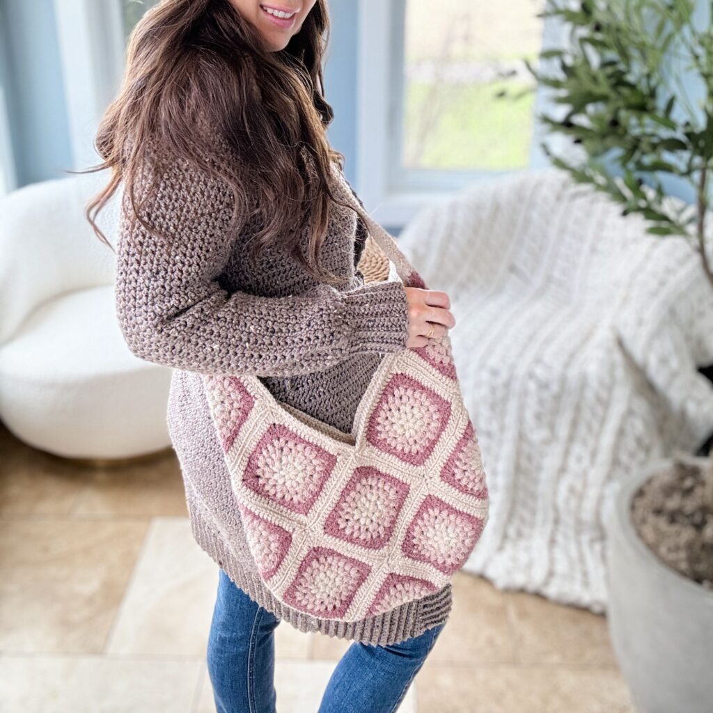 A smiling woman with long brown hair, wearing a chunky knit sweater, holds a large crocheted purse with a geometric pattern.