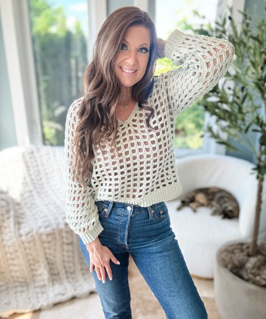 A person with long brown hair wearing a white crochet mesh top and jeans poses in a bright room, with a cat resting on a cream-colored chair in the background.