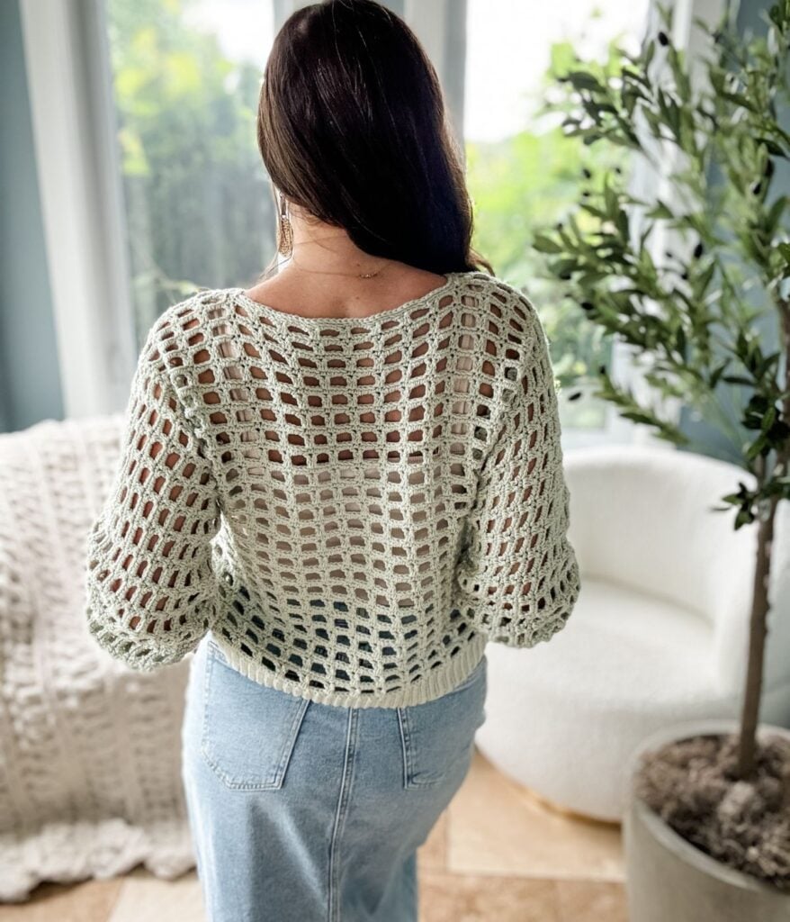 Person with long brown hair facing away, wearing a light green crochet mesh top and blue jeans, standing in a room with a white chair and potted plant next to a window.