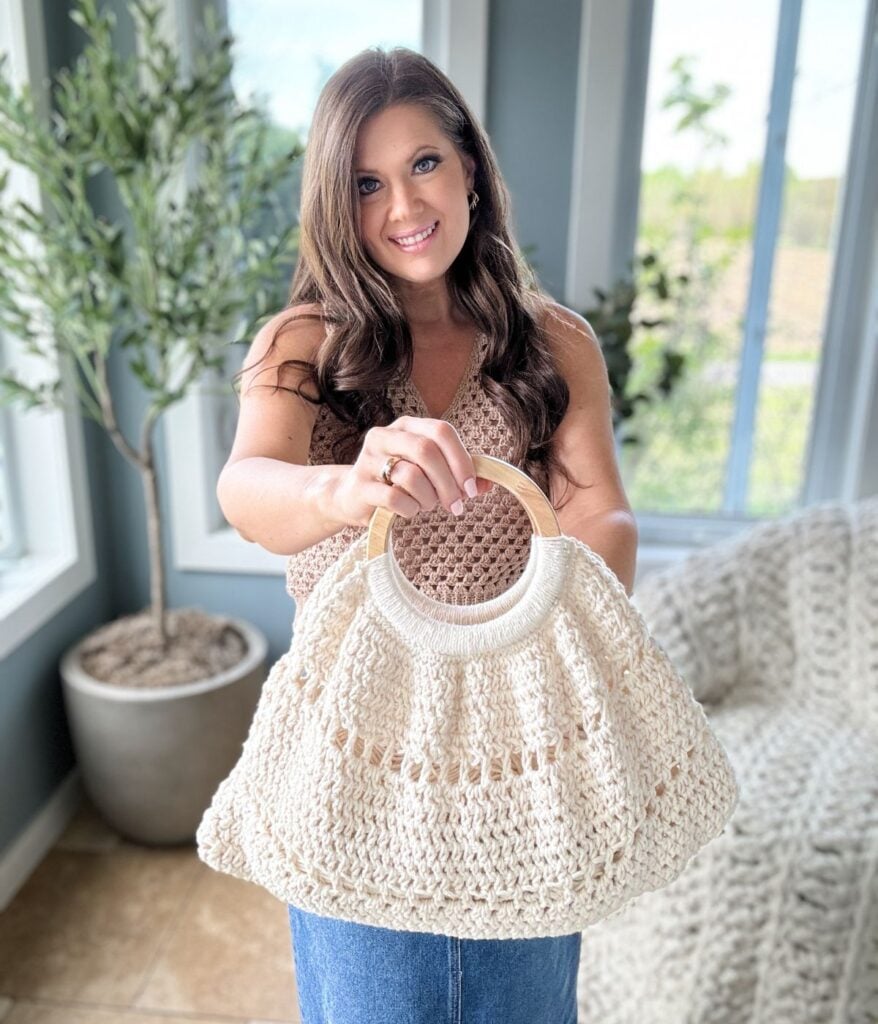 A woman with long hair smiling and holding a cream-colored crochet purse inside a room with plants and a window.