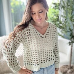 A woman with long brown hair wearing a light green crochet mesh top and jeans is standing indoors near a plant.