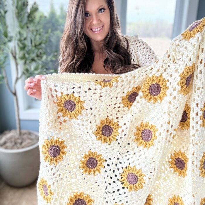 A woman with long, wavy brown hair smiling and holding up a handmade crochet blanket with a yellow and brown sunflower design.