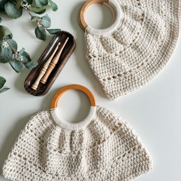 A flat lay of a cream crocheted bag and matching hat with wooden handles, featuring a free pattern, accompanied by a dark case with makeup brushes, set against a light background with green leaves.