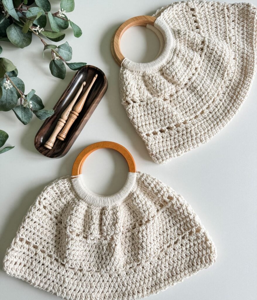 A flat lay of a cream crocheted bag and matching hat with wooden handles, featuring a free pattern, accompanied by a dark case with makeup brushes, set against a light background with green leaves.