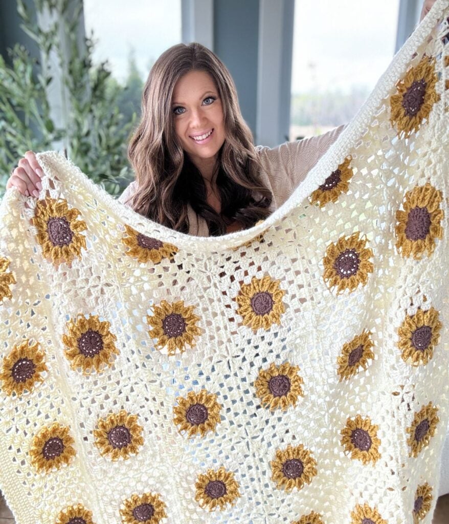 A woman with brown hair smiles while holding up a large, handmade blanket with a sunflower pattern.