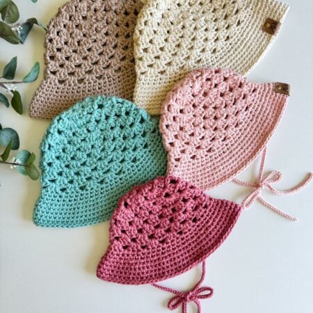 Five crocheted hats in various colors (brown, beige, pink, teal, and red) arranged side by side on a white surface, with a few sprigs of greenery nearby. One hat features an intricate crochet top detail.