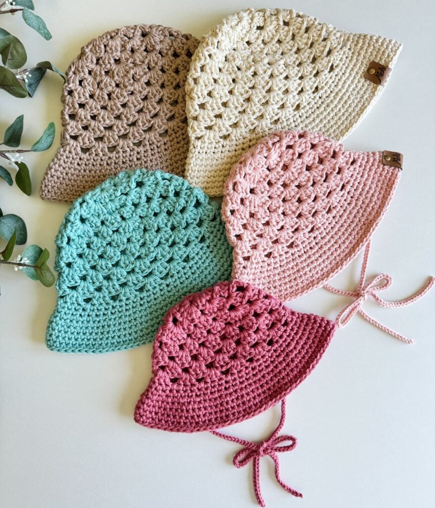 Five crocheted hats in various colors (brown, beige, pink, teal, and red) arranged side by side on a white surface, with a few sprigs of greenery nearby. One hat features an intricate crochet top detail.