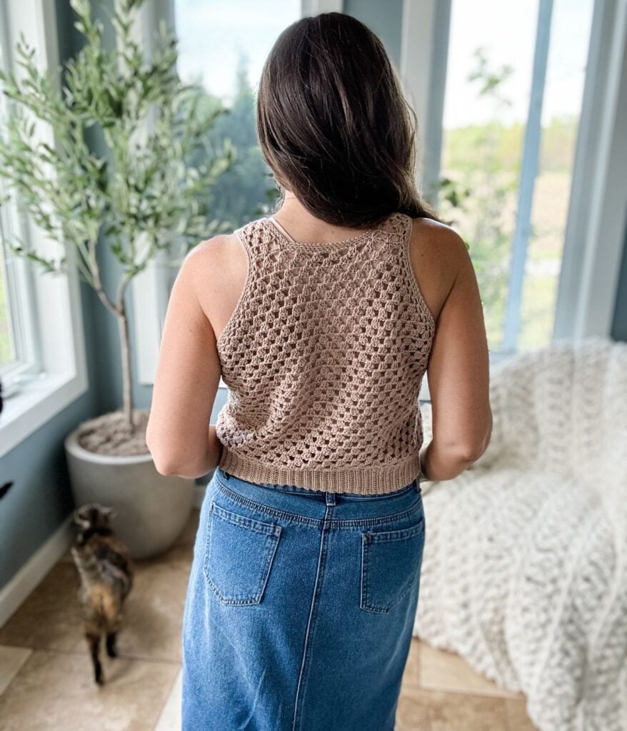 A person with long hair, wearing a sleeveless crochet tank top made from a free crochet pattern and a denim skirt, stands indoors with their back to the camera. A small cat is near their feet.