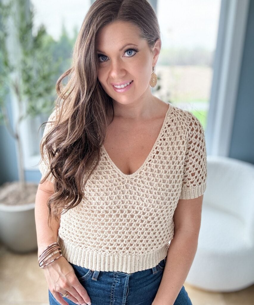 A woman with long brown hair and blue eyes is wearing a cream-colored crochet top and blue jeans, standing indoors with a plant and chair in the background.