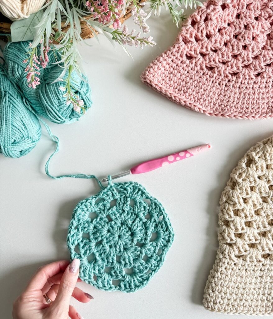 A hand holding a blue crocheted doily in progress with a pink crochet hook, surrounded by balls of yarn, finished crocheted hats, and a colorful crochet top in the making.