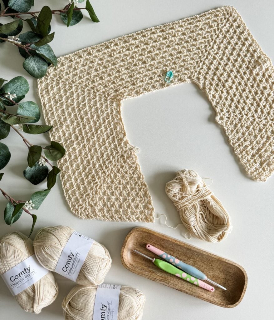 A partially crocheted beige fabric with a geometric pattern, surrounded by yarn and needles in a wooden tray, hints at the beginnings of a beautiful crochet top. Eucalyptus leaves placed on the white surface add a touch of nature to this serene crafting scene.