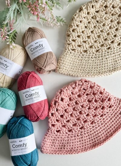 Two crochet hats made of beige and pink yarn are displayed alongside five skeins of various colored yarn, a crochet top, and some foliage on a white surface.