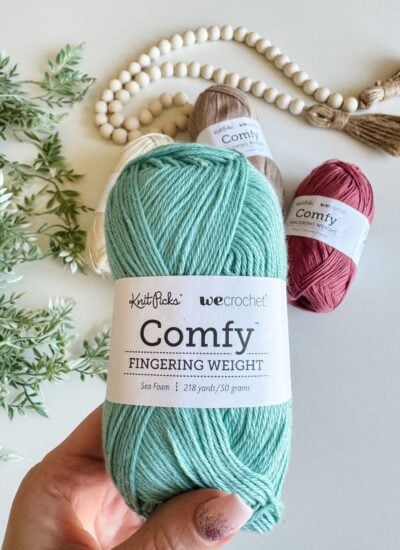 A hand holds a skein of sea foam green yarn labeled "Knit Picks Wee Crochet Comfy Fingering Weight," perfect for crafting a delicate crochet top. Other skeins in beige and pink, a bead garland, and green plant leaves are in the background.
