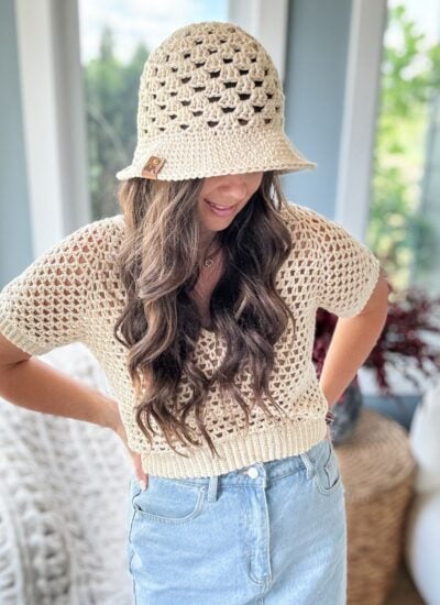 A woman with long hair wears a beige crochet top and matching hat, paired with a light blue denim skirt. She stands indoors near a window, her hands resting on her hips.